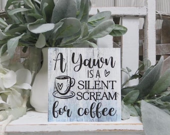 May include: A white wooden sign with blue and grey paint. The sign reads "A yawn is a silent scream for coffee" with a small heart and a coffee cup with a heart on it.