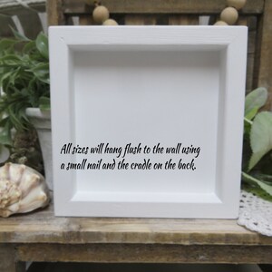 May include: White square frame with a white mat and a black inscription that reads: "All sizes will hang flush to the wall using a small nail and the cradle on the back."