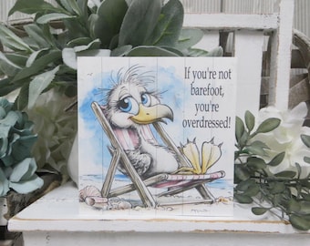 May include: A square wooden sign featuring a cartoon seagull with large blue eyes sitting in a beach chair. The text reads, "If you're not barefoot, you're overdressed!" The illustration includes a sandy beach background with seashells and blue watercolor accents.