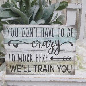 Peut inclure: Une pancarte en bois avec un fond blanc vieilli et un grain de bois peint en bleu et vert. La pancarte indique "You don't have to be crazy to work here, we'll train you".