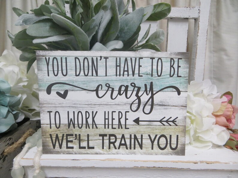 Peut inclure: Une pancarte en bois avec un fond blanc vieilli et un grain de bois peint en bleu et vert. La pancarte indique "You don't have to be crazy to work here, we'll train you".