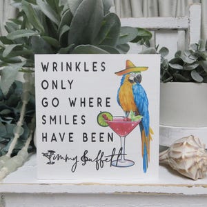 May include: A white sign with black text that reads "Wrinkles only go where smiles have been" with a drawing of a parrot wearing a sombrero and holding a pink cocktail. The text "Jimmy Buffett" is written below the quote.