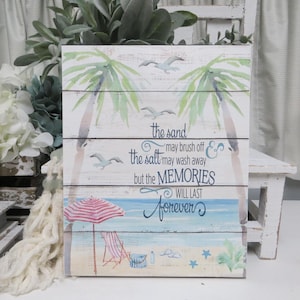 May include: A white wooden sign with a beach scene and the text "the sand may brush off & the salt may wash away but the MEMORIES WILL LAST forever".