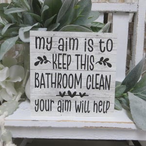 May include: White wooden sign with black text that reads "My aim is to keep this bathroom clean. Your aim will help."