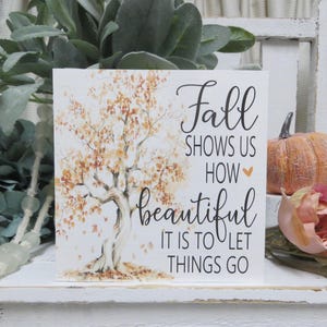 May include: A square, white sign with the text "Fall shows us how beautiful it is to let things go." The sign features a watercolor illustration of a tree with orange and brown leaves. A small pumpkin and a rose are in the background.