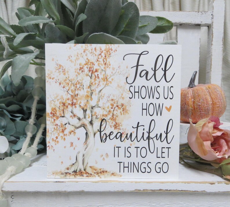 May include: A square, white sign with the text "Fall shows us how beautiful it is to let things go." The sign features a watercolor illustration of a tree with orange and brown leaves. A small pumpkin and a rose are in the background.