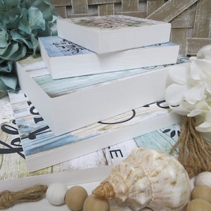 May include: A stack of four white-framed wooden signs with various designs. The signs feature floral, script, and wood grain patterns in blue, white, and black. Decorative elements include a seashell, wooden beads, and faux flowers.