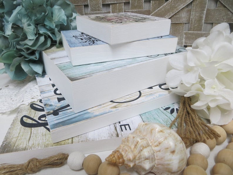 May include: A stack of four white-framed wooden signs with various designs. The signs feature floral, script, and wood grain patterns in blue, white, and black. Decorative elements include a seashell, wooden beads, and faux flowers.