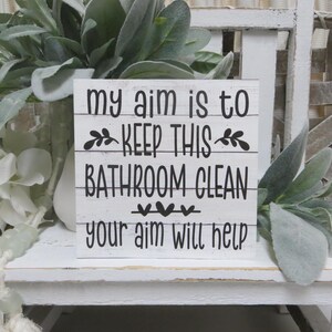 May include: White wooden sign with black text that reads "My aim is to keep this bathroom clean. Your aim will help."