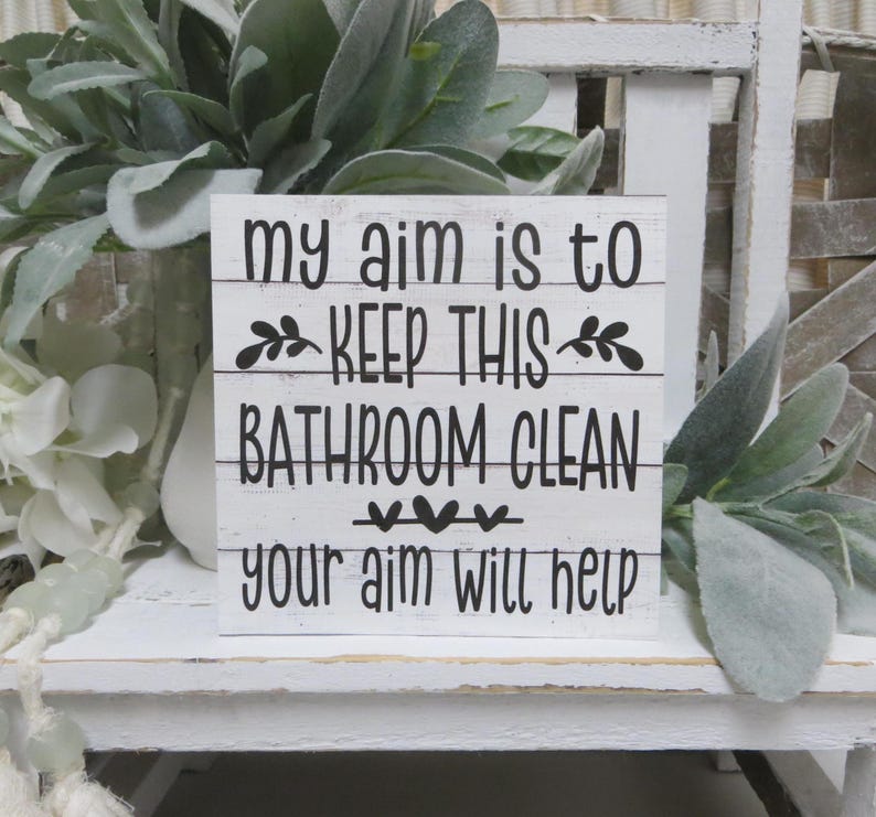 May include: White wooden sign with black text that reads "My aim is to keep this bathroom clean. Your aim will help."