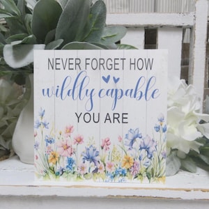 May include: A white wooden sign with the text "NEVER FORGET HOW wildly capable YOU ARE" in blue and black. The bottom of the sign features a watercolor floral design with pink, blue, and yellow flowers.
