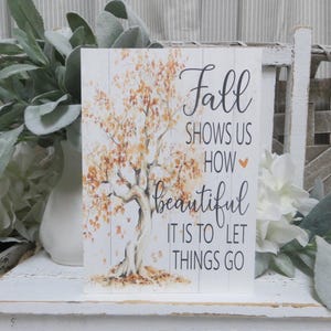 May include: A rectangular wooden sign with a white background and a tree illustration with orange and yellow leaves. The sign has the words "Fall shows us how beautiful it is to let things go" in black script.