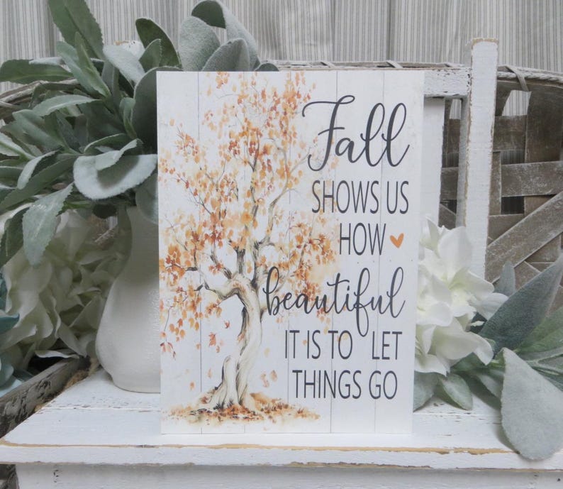 May include: A rectangular wooden sign with a white background and a tree illustration with orange and yellow leaves. The sign has the words "Fall shows us how beautiful it is to let things go" in black script.