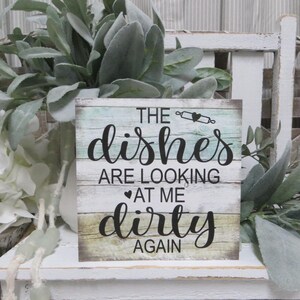 May include: A rustic wooden sign with a distressed whitewash finish. The sign features a teal and white background with the text "The dishes are looking at me dirty again".