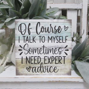 May include: A wooden sign with a light blue and green background and white wood grain texture. The sign reads "Of Course I Talk To Myself Sometimes I Need Expert Advice" with a heart symbol before and after the word "advice".