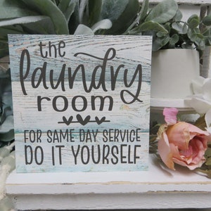 May include: A wooden sign with a blue and white distressed paint finish. The sign reads "The Laundry Room For Same Day Service Do It Yourself" with three small hearts above the text.