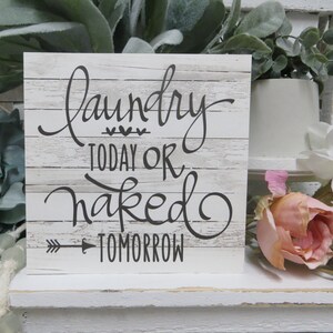 May include: White wooden sign with black text that reads "Laundry Today or Naked Tomorrow" with a small arrow pointing to the right. The sign is sitting on a white shelf with green and pink flowers.