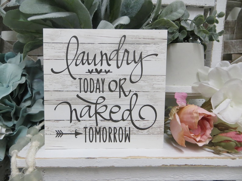 May include: White wooden sign with black text that reads "Laundry Today or Naked Tomorrow" with a small arrow pointing to the right. The sign is sitting on a white shelf with green and pink flowers.