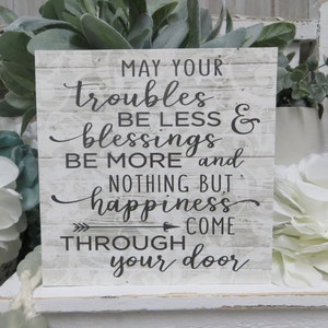 May include: White wooden sign with black text that reads "May your troubles be less & blessings be more and nothing but happiness come through your door".
