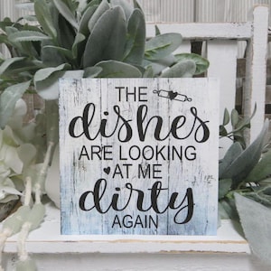 May include: White wooden sign with blue and white distressed paint. The sign says "The dishes are looking at me dirty again".
