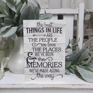 May include: White wooden sign with black text that reads "the best THINGS IN LIFE ARE THE PEOPLE we love THE PLACES WE'VE BEEN & the MEMORIES WE'VE MADE ALONG the way"