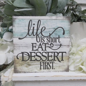 May include: A rustic wooden sign with a blue and green background. The sign reads "Life is short eat dessert first."