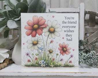May include: A square greeting card featuring a watercolor illustration of pink and white daisies with green stems and leaves. The text on the right side reads, You're the friend everyone wishes they had, accompanied by small pink hearts and bubbles.