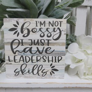 May include: A white wooden sign with a distressed woodgrain finish. The sign reads "I'm not bossy I just have leadership skills" in black script font with a floral design at the top and bottom.