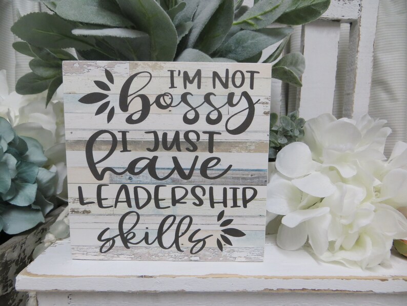 May include: A white wooden sign with a distressed woodgrain finish. The sign reads "I'm not bossy I just have leadership skills" in black script font with a floral design at the top and bottom.