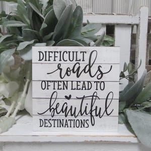 May include: White wooden sign with black text that reads "Difficult roads often lead to beautiful destinations".