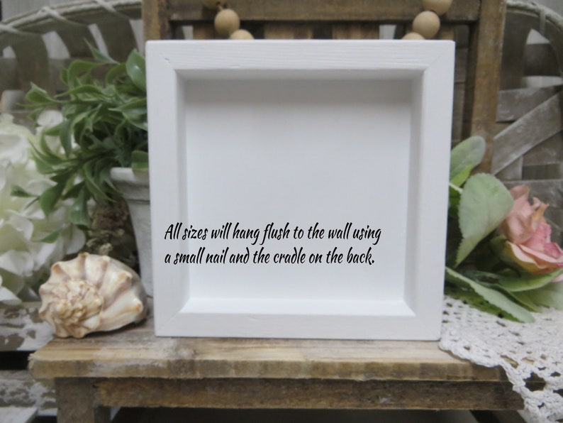 May include: White square frame with a white mat and a black inscription that reads: "All sizes will hang flush to the wall using a small nail and the cradle on the back."