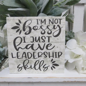 May include: A white wooden sign with black text that reads "I'm not bossy I just have leadership skills". The sign has a distressed wood look and is decorated with small black floral designs.