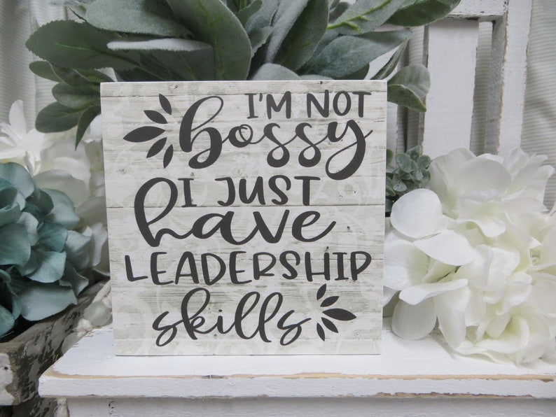 May include: A white wooden sign with black text that reads "I'm not bossy I just have leadership skills". The sign has a distressed wood look and is decorated with small black floral designs.