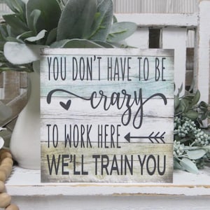 May include: A square wooden sign with a weathered, multi-colored wood grain finish in shades of blue, green, and white. The sign features black text that reads, "YOU DON'T HAVE TO BE crazy TO WORK HERE WE'LL TRAIN YOU," with a small heart and arrow.