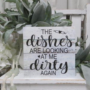May include: A white wooden sign with blue and green painted stripes. The sign reads "The dishes are looking at me dirty again".