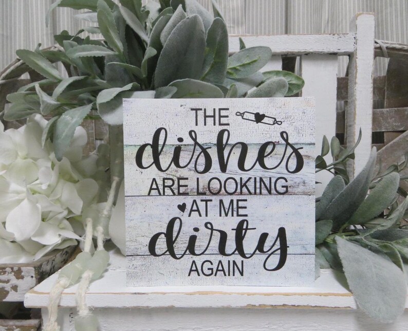 May include: A white wooden sign with blue and green painted stripes. The sign reads "The dishes are looking at me dirty again".