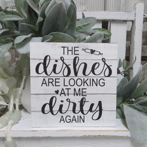 May include: White wooden sign with black text that reads "The dishes are looking at me dirty again". The sign has a distressed look and is decorated with a small heart and a rolling pin.