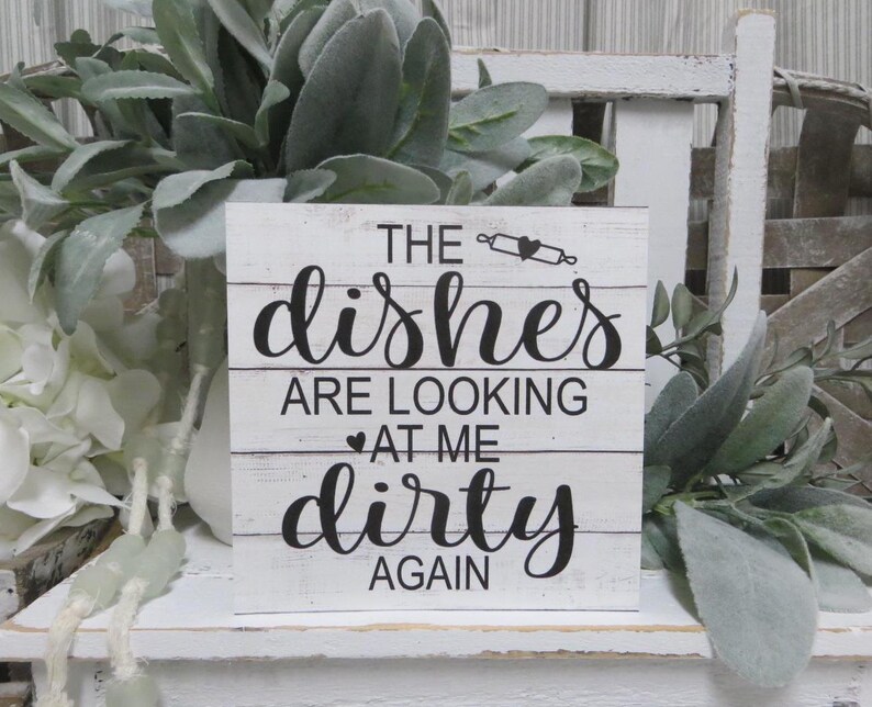 May include: White wooden sign with black text that reads "The dishes are looking at me dirty again". The sign has a distressed look and is decorated with a small heart and a rolling pin.