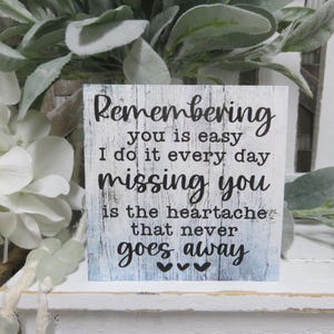 May include: White wooden sign with black text that reads "Remembering you is easy I do it every day missing you is the heartache that never goes away".