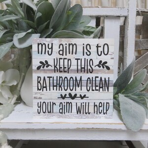 May include: White wooden sign with the text "My aim is to keep this bathroom clean your aim will help". The sign has a distressed wood look and is decorated with small black hearts.
