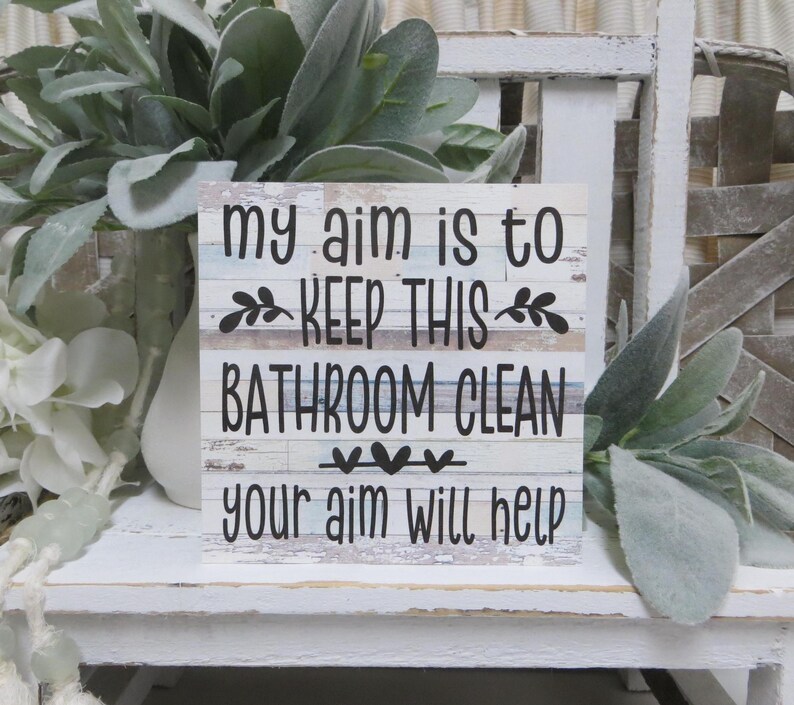 May include: White wooden sign with the text "My aim is to keep this bathroom clean your aim will help". The sign has a distressed wood look and is decorated with small black hearts.