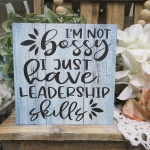 May include: A blue wooden sign with white painted wood grain and black lettering. The sign reads "I'm not bossy I just have leadership skills".
