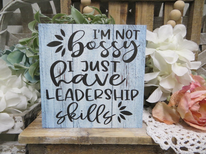 May include: A blue wooden sign with white painted wood grain and black lettering. The sign reads "I'm not bossy I just have leadership skills".