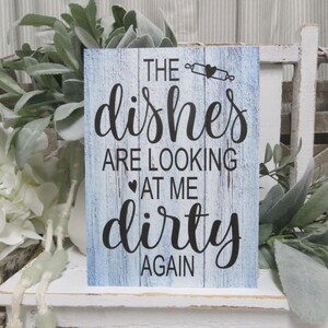 May include: A blue wooden sign with white distressed paint and black lettering that reads "The dishes are looking at me dirty again".