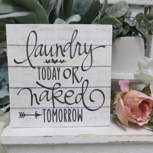 May include: White wooden sign with black lettering that reads "Laundry Today or Naked Tomorrow" with a small arrow pointing to the right. The sign is sitting on a white shelf with green plants and pink flowers.