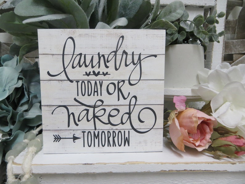 May include: White wooden sign with black lettering that reads "Laundry Today or Naked Tomorrow" with a small arrow pointing to the right. The sign is sitting on a white shelf with green plants and pink flowers.
