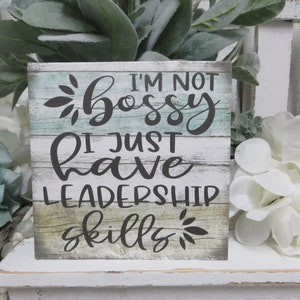 May include: A wooden sign with a distressed white background and a teal blue stripe. The sign reads "I'm not bossy I just have leadership skills" in black cursive font with a sunburst design at the top and bottom.