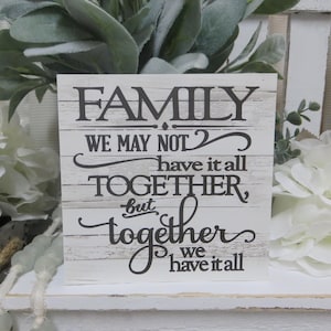 May include: White wooden sign with black text that reads "FAMILY WE MAY NOT have it all TOGETHER, but together WE have it all".