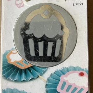 May include: A cupcake-shaped paper punch with the words "large punch" in a package. The punch is silver and gold. Below are blue paper fans with cupcake cutouts in pink and white.