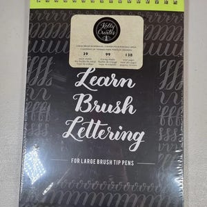 May include: A spiral-bound workbook for brush lettering. The cover is black with white lettering that reads "Learn Brush Lettering." The workbook includes 39 work sheets, 99 tracing sheets, and 138 total pages. The top edge is neon yellow.